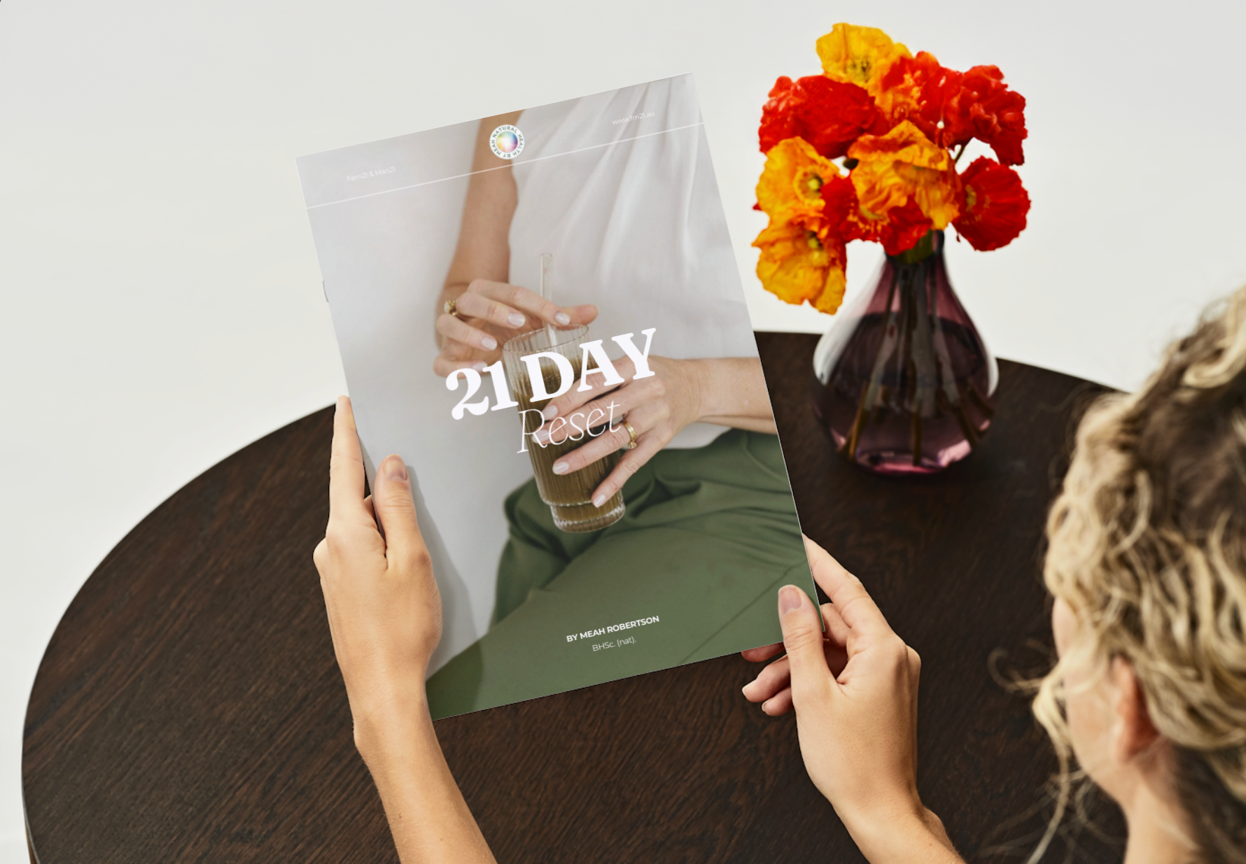 Person holding a '21 Day' book with a vase of flowers on a table