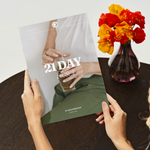 Person holding a '21 Day' book with a vase of flowers on a table